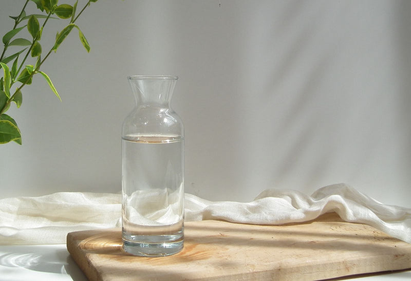 Carafe de verre et linge blanc sur une planche de bois clair, le circuit remis au propre avant de changer de senteur dans un diffuseur sans mélanger les parfums